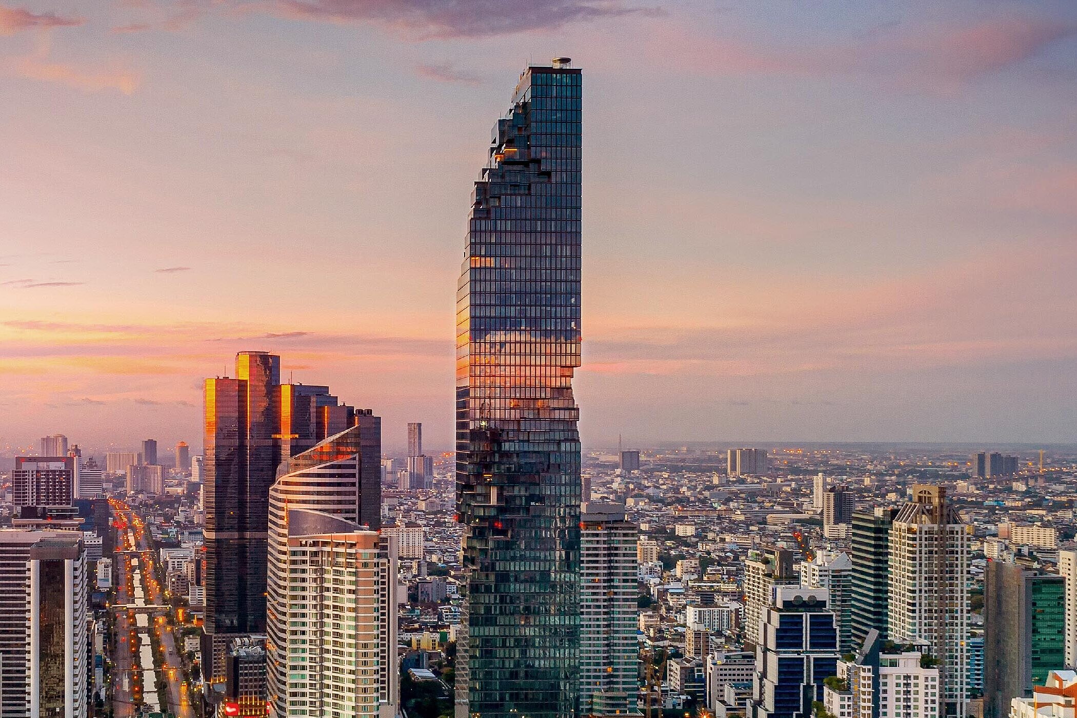 Eine atemberaubende Skyline einer Stadt bei Sonnenuntergang. Hohe, moderne Wolkenkratzer dominieren das Bild, während die sanften Farben des Himmels orange und lila Nuancen zeigen. Die Architektur ist vielfältig, mit reflektierenden Glasfassaden und einzigartigen Formen.