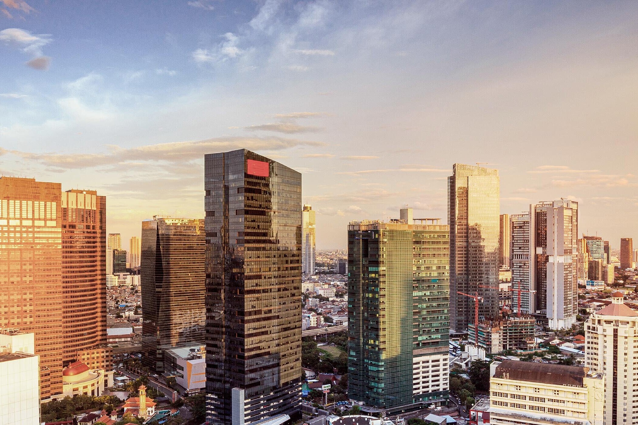 Eine Panoramaansicht einer modernen Stadt mit hohen Glas- und Stahlgebäuden, die im Licht der untergehenden Sonne funkeln. Der Himmel ist in sanften Farben gefärbt, während Straßen und Grünflächen sichtbar sind.