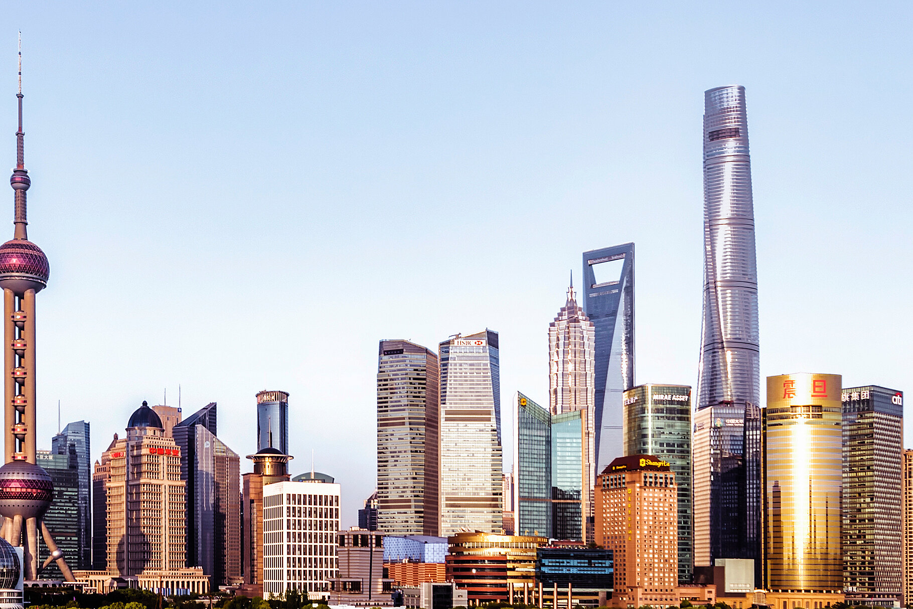 Panoramaaufnahme der Skyline von Shanghai bei Sonnenaufgang, mit markanten Wolkenkratzern wie dem Oriental Pearl Tower und dem Shanghai Tower, die das Stadtbild prägen. Der Vordergrund zeigt das Wasser des Flusses, der entlang der Stadt fließt.