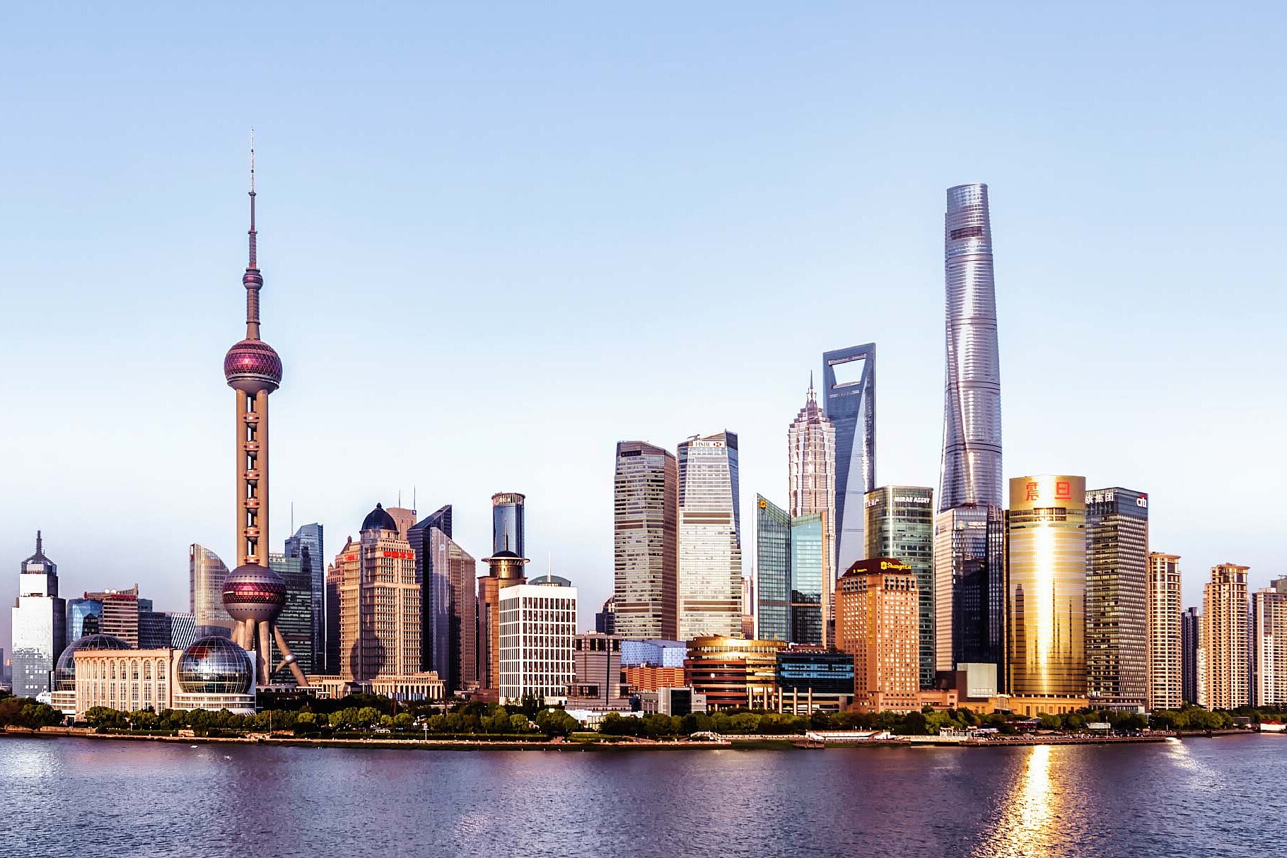 Skyline von Shanghai, mit modernen Wolkenkratzern und dem ikonischen Oriental Pearl Tower in der Mitte. Im Vordergrund ist der Fluss zu sehen, mit einigen Booten, die vorbeifahren. Der Himmel ist klar und strahlend.