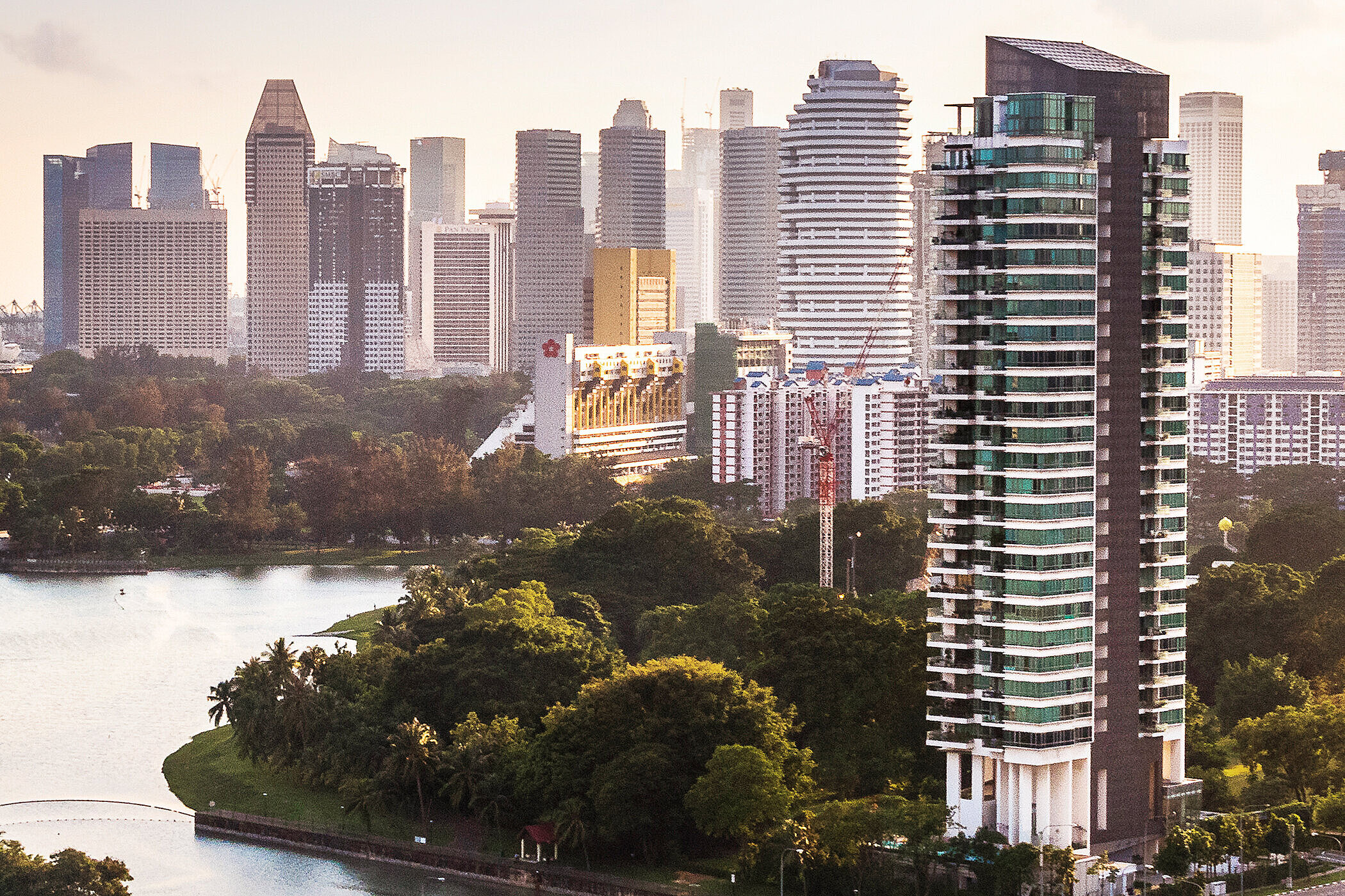 Blick auf eine moderne Stadtlandschaft mit hohen Gebäuden, umgeben von Grünflächen, einem Fluss und einer Promenade. Im Hintergrund ist ein Riesenrad und eine Uferpromenade zu sehen. Die Szene vermittelt eine Mischung aus Natur und urbanem Leben im frühen Morgenlicht.