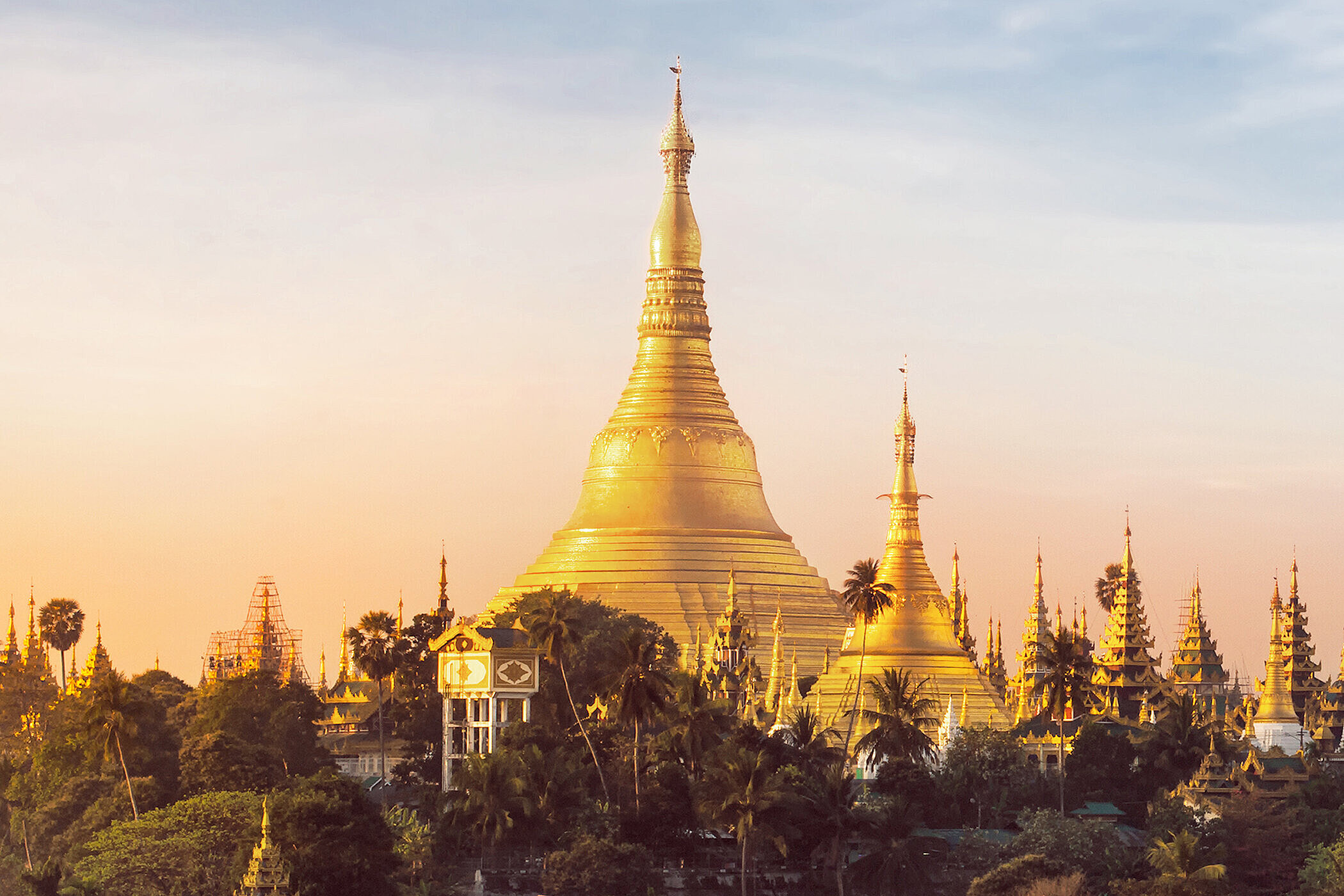 Eine goldene Stupa erhebt sich majestätisch vor einem sanften Sonnenuntergang. Umgeben von weiteren Pagoden und üppigem Grün, strahlt die Architektur in warmen Farben und vermittelt eine friedliche Atmosphäre. Die Szene symbolisiert die spirituelle und kulturelle Bedeutung des Ortes.