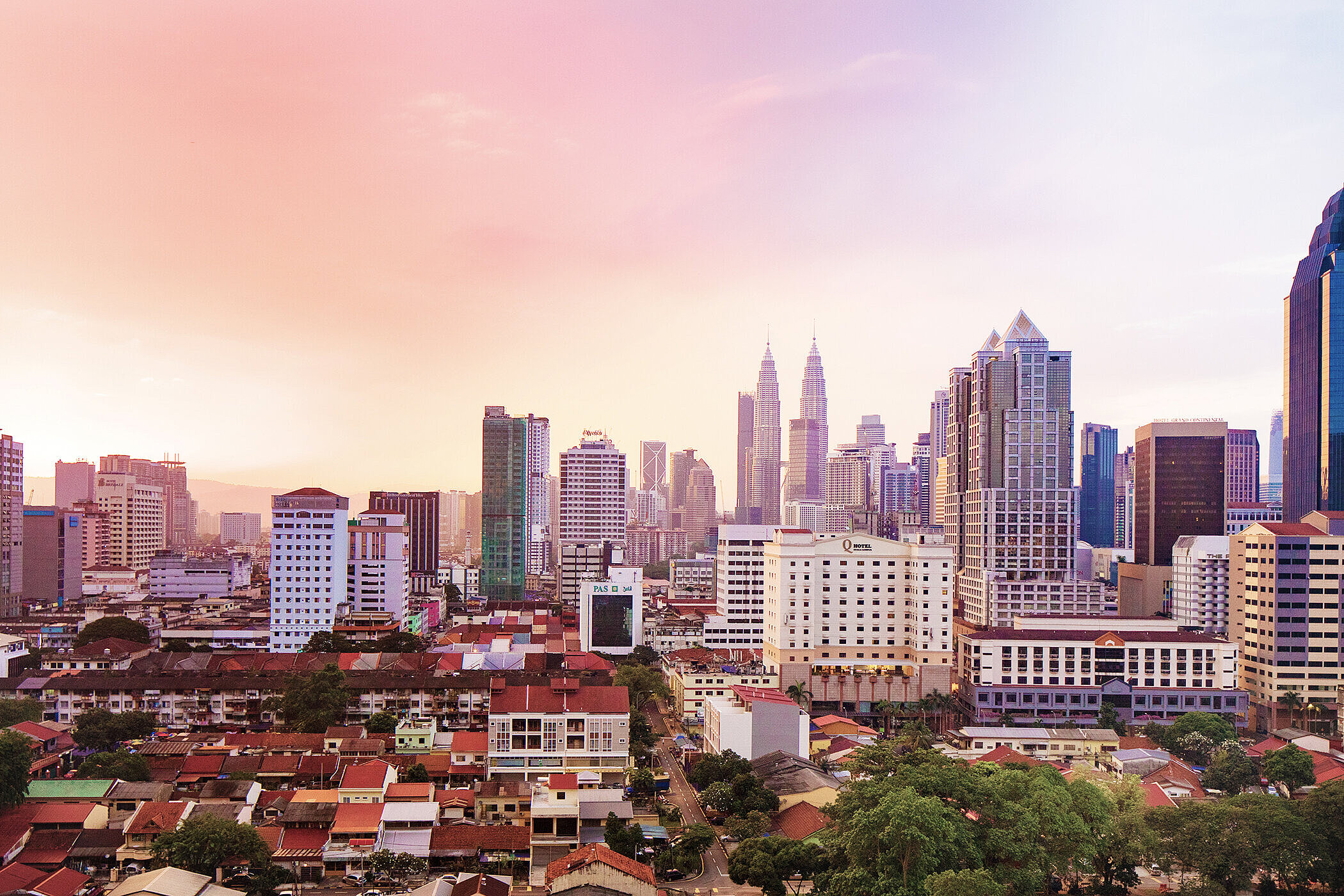Die Skyline einer Stadt mit modernen Wolkenkratzern und einem sanften Sonnenuntergang im Hintergrund. Vorne sind niedrigere Gebäude und grüne Bäume zu sehen. Die Mischung aus Architektur und Natur schafft eine dynamische städtische Atmosphäre.