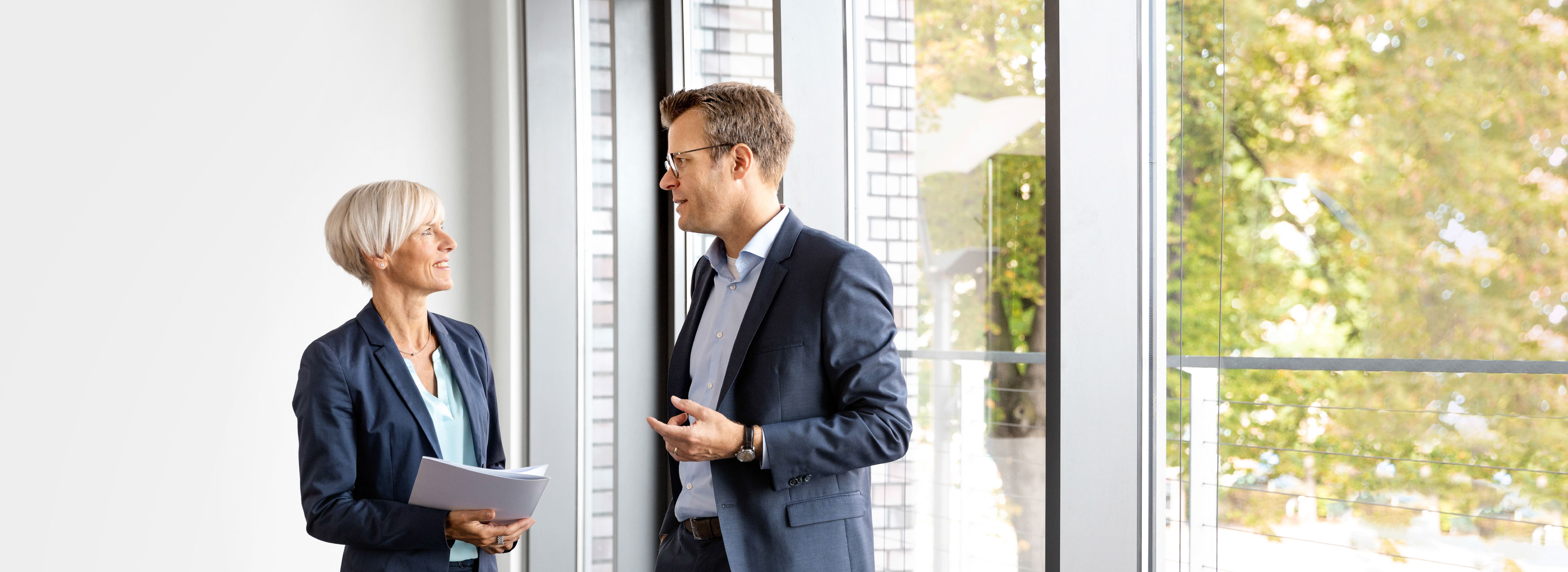 Ein Mann und eine Frau stehen in einem modernen Büro und führen ein Gespräch. Beide sind formell gekleidet, der Mann hat eine Brille. Im Hintergrund sind große Fenster, durch die die Bäume im Freien sichtbar sind.