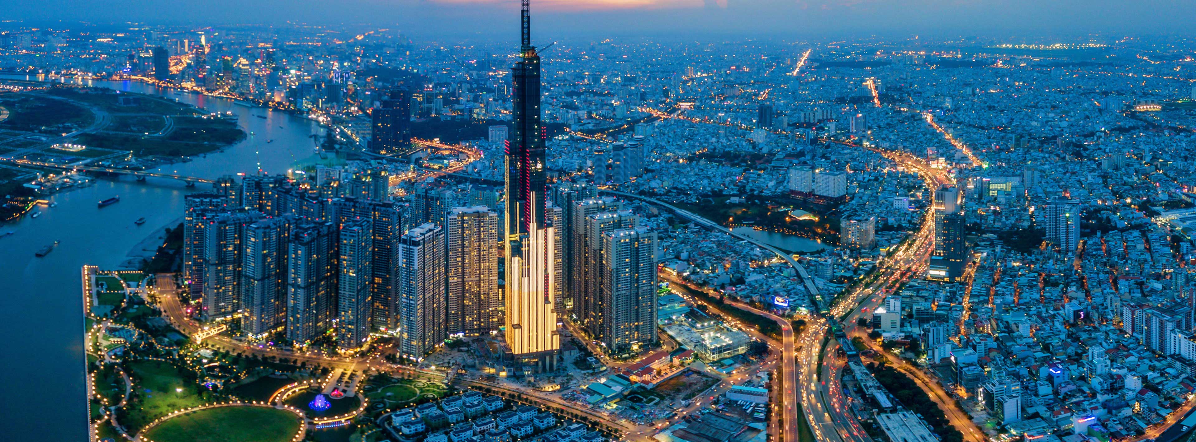 Die Aufnahme zeigt eine beeindruckende Skyline mit dem höchsten Gebäude der Stadt, das hell erleuchtet ist. Umgeben von vielen weiteren Hochhäusern und einem Fluss im Vordergrund, strahlt die Stadt bei Dämmerung lebendige Lichter aus.