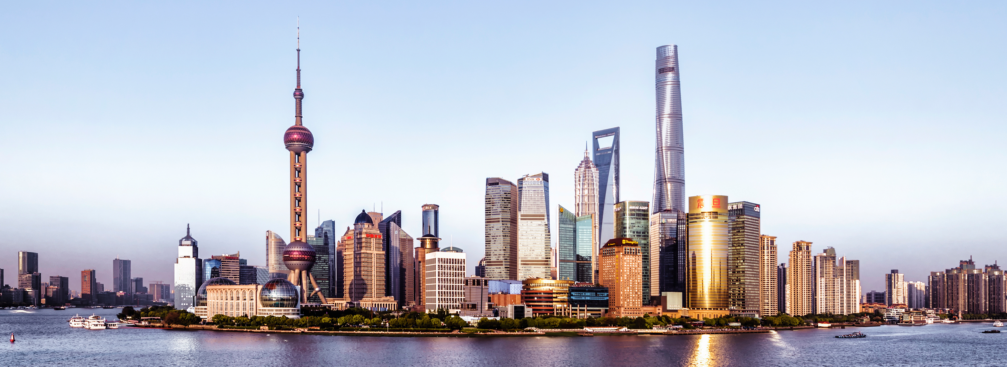 Blick auf die Skyline von Shanghai mit modernen Wolkenkratzern, darunter der markante Shanghai Tower und der Oriental Pearl Tower. Der Huangpu-Fluss fließt im Vordergrund, umrahmt von Gebäuden und Bäumen. Der Himmel ist klar und hell, was die beeindruckende Architektur zur Geltung bringt.