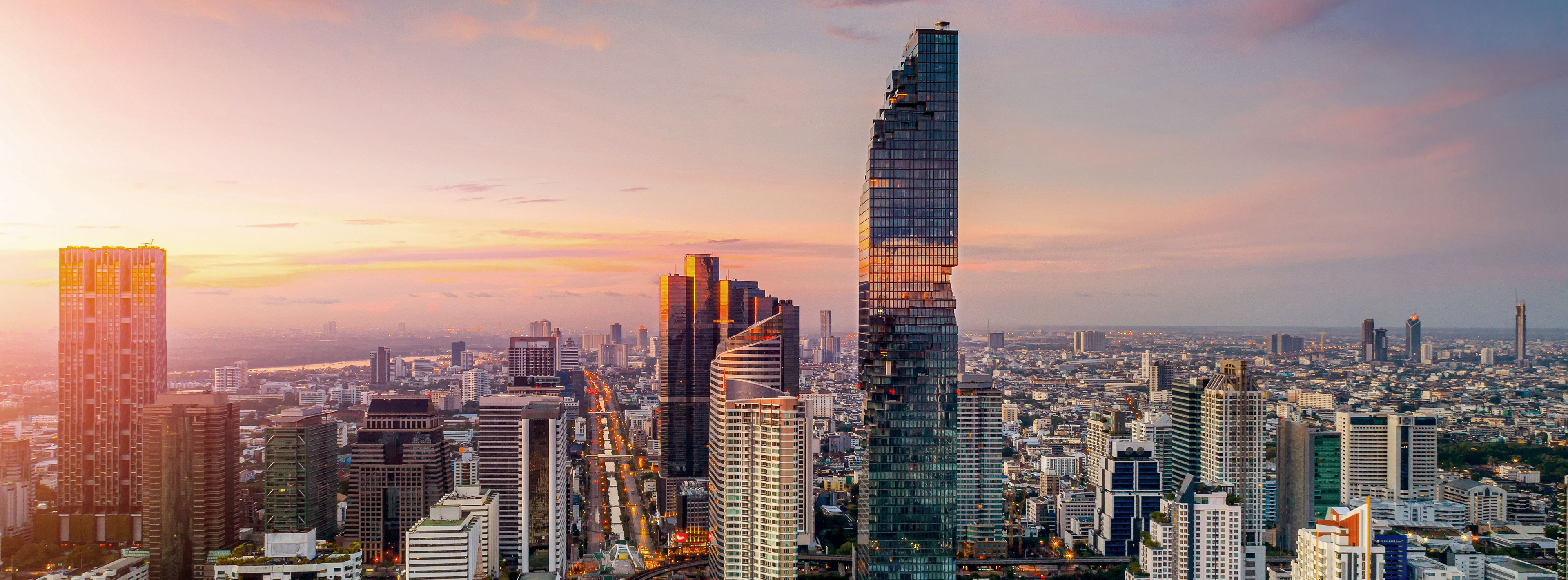 Die Skyline einer modernen Stadt wird bei Sonnenuntergang dargestellt. Hohe Wolkenkratzer, darunter ein markanter gläserner Turm, sind sichtbar. Der Himmel zeigt sanfte Farbverläufe in Orange und Lila, die der Szene eine ruhige Atmosphäre verleihen. Die Stadt breitet sich weit unter dem Himmel aus.