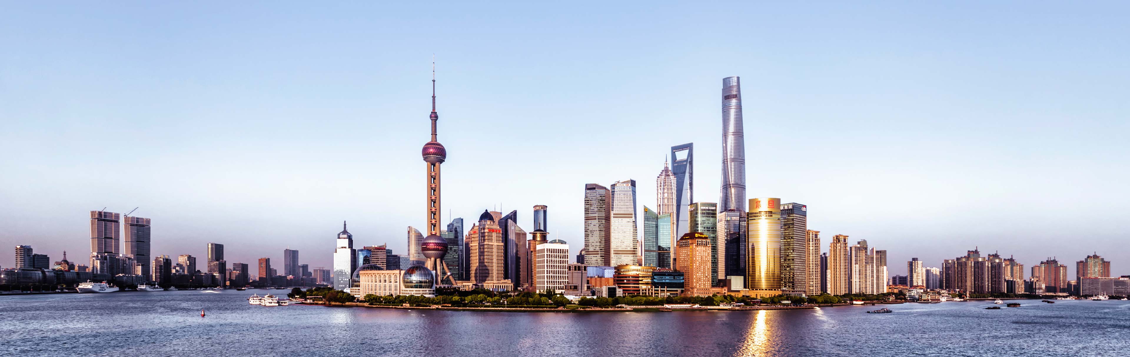 Skyline von Shanghai, mit modernen Wolkenkratzern und dem ikonischen Oriental Pearl Tower in der Mitte. Im Vordergrund ist der Fluss zu sehen, mit einigen Booten, die vorbeifahren. Der Himmel ist klar und strahlend.