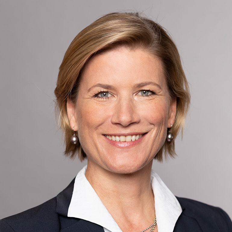 Ein Porträt einer Frau mit kurzen blonden Haaren, die ein freundliches Lächeln zeigt. Sie trägt einen dunklen Blazer und eine weiße Bluse. Der Hintergrund ist einfarbig grau, was den Fokus auf ihr Gesicht lenkt.